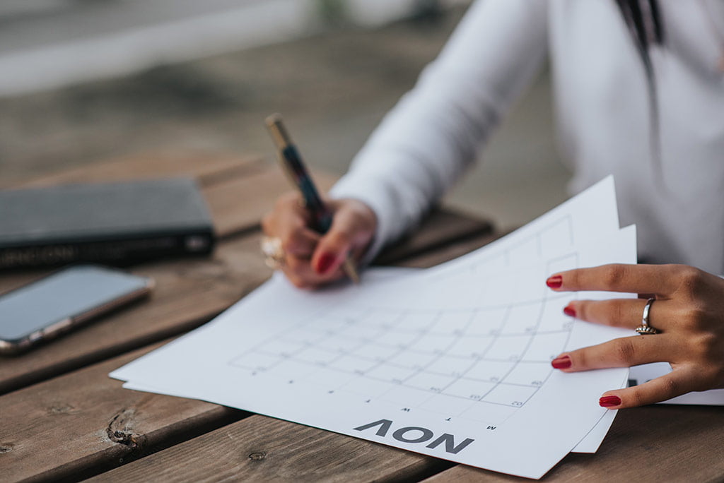 Como fazer um calend&aacute;rio de conte&uacute;do para Instagram? : mulher preenchendo folha de calendario com uma caneta em cima de uma mesa de madeira