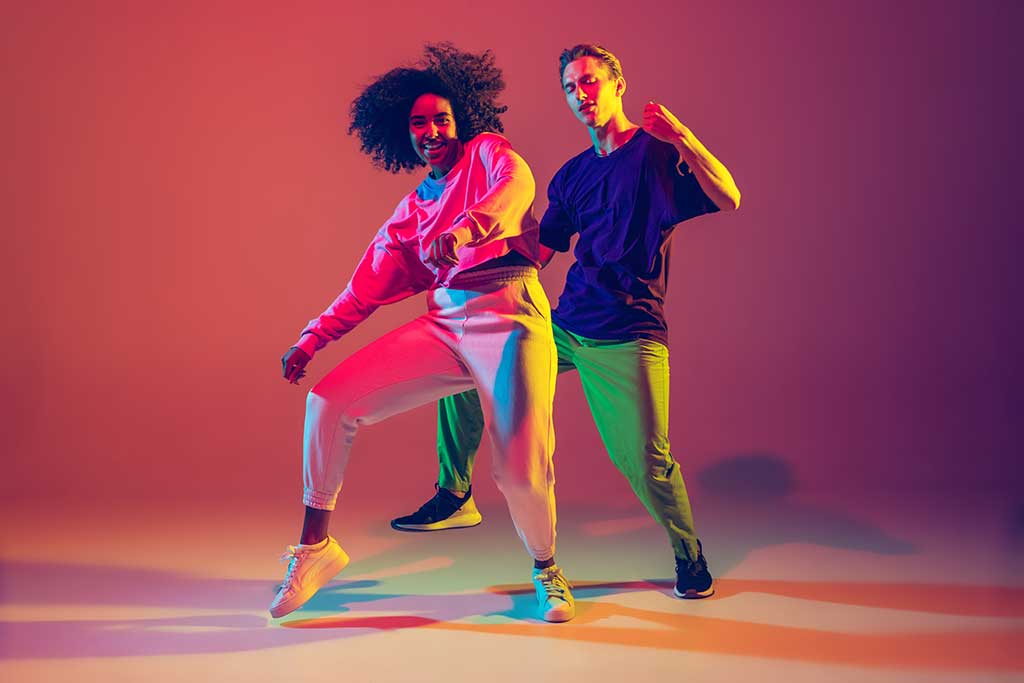 Conhe&ccedil;a as microtrends do Tik Tok : Dupla dan&ccedil;ando com roupas coloridas em um fundo avermelhado