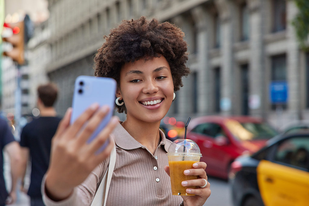 O que &eacute; um nano influencer? : influenciadora com um copo de uma bebida amarelada fazendo grava&ccedil;&atilde;o com um celular em plena rua cheia de carros
