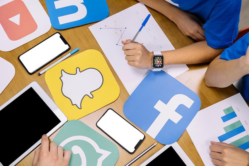 Tipos de conte&uacute;do digital : redes sociais : V&aacute;rias logos de redes sociais impressas em cima de uma mesa com canetas, tablets, celulare e m&atilde;os nelas