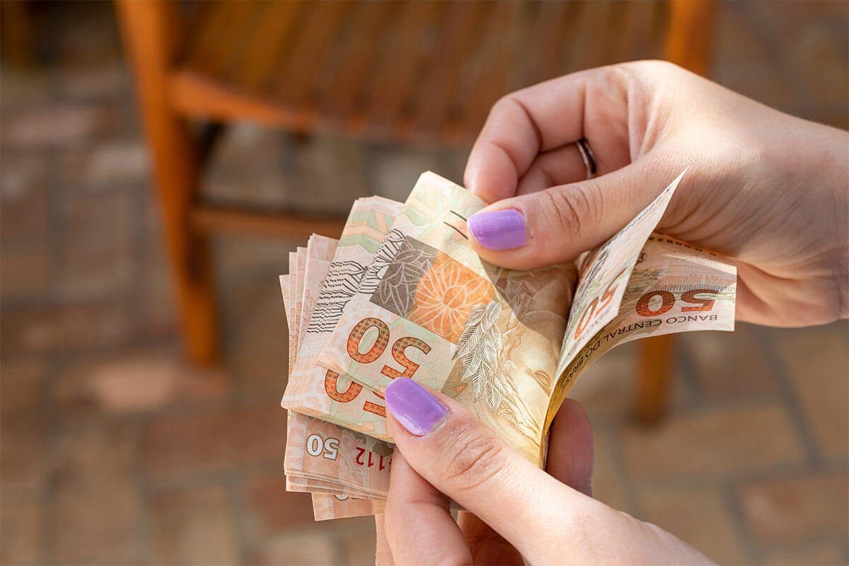 M&atilde;os femininas contando v&aacute;rias notas de cinquenta reais, fazendo refer&ecirc;ncia ao valor que um influencer ganha. Foto: @Freepik
