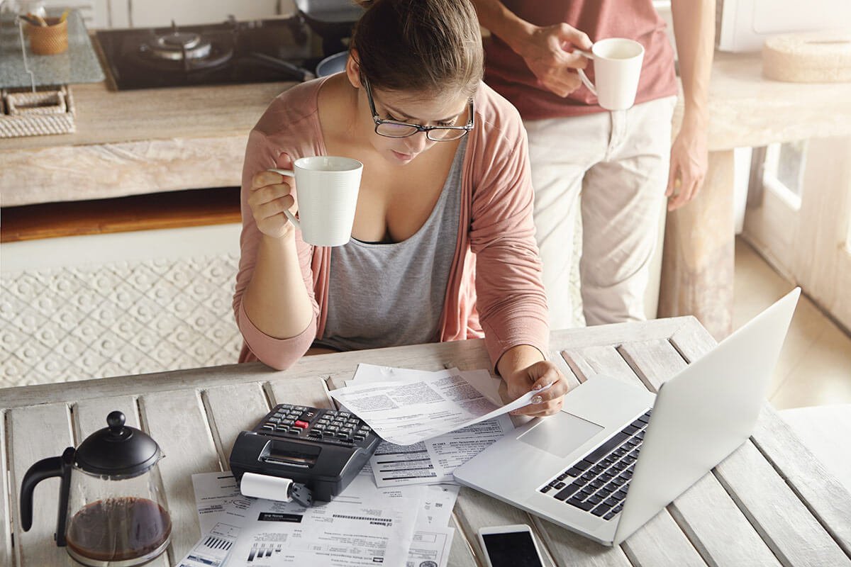 Influencer de &oacute;culos com uma x&iacute;cara na m&atilde;o, observando pap&eacute;is na mesa e ao lado, uma calculadora, um celular, um notebook e uma garrafa de caf&eacute;, sugerindo an&aacute;lise dos impostos referentes ao MEI. Foto: @Freepik