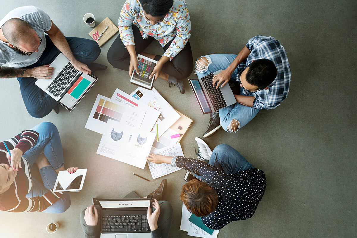 Equipe de trabalho sentada no ch&atilde;o, com computadores, tablets, pap&eacute;is e copos de caf&eacute;, trabalhando juntos para manter uma melhor integra&ccedil;&atilde;o. Foto: @Freepik.
