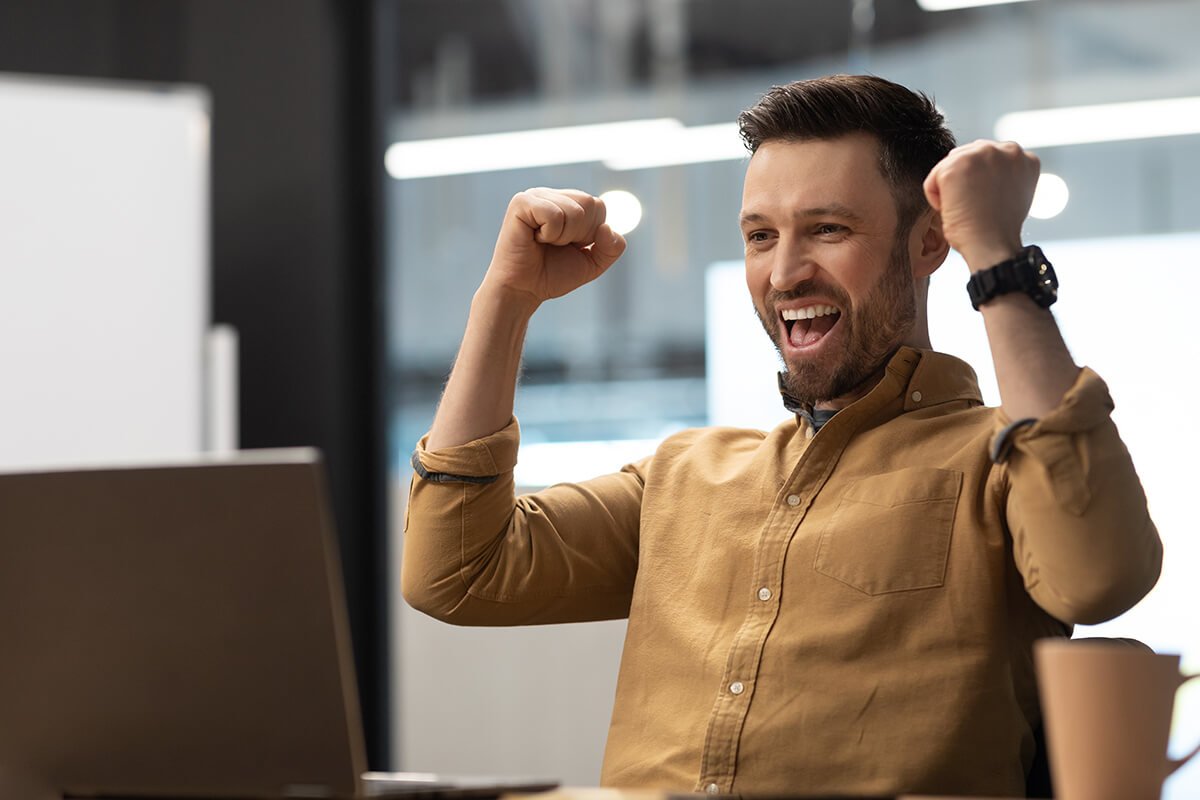 Um homem diante de um notebook, comemorando uma vit&oacute;ria com os bra&ccedil;os para cima, ilustrando a for&ccedil;a motivadora da gamifica&ccedil;&atilde;o. Foto: @Freepik.