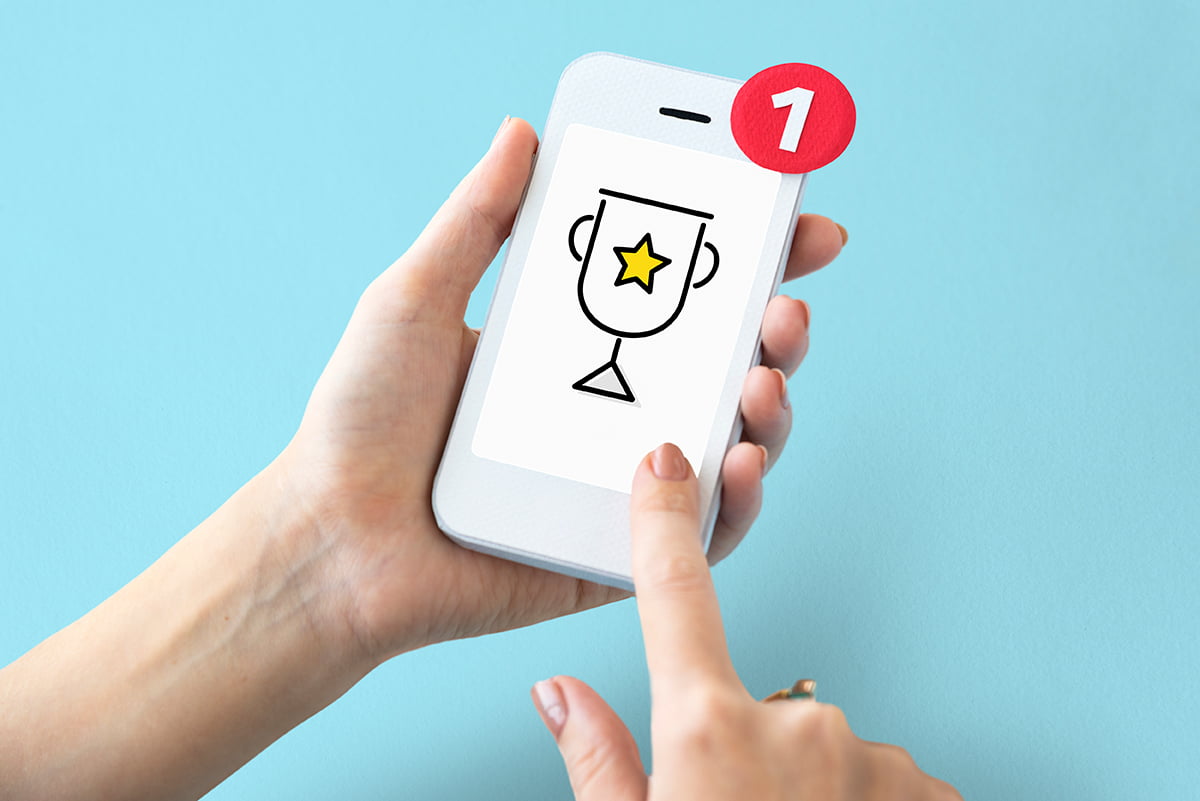 M&atilde;os segurando um celular com a imagem de um trof&eacute;u, representando um dos benef&iacute;cios da gamifica&ccedil;&atilde;o que &eacute; o aumento de engajamento. Foto: @Freepik.