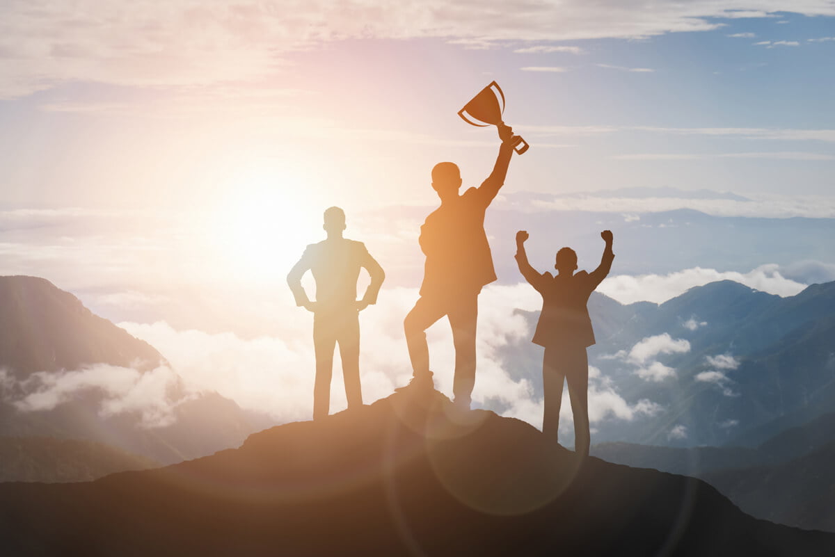 Imagem de tr&ecirc;s pessoas no topo da montanha, sendo uma delas com um trof&eacute;u levantado e outra com as m&atilde;os para cima, representando a recompensa pelo sucesso nas estrat&eacute;gias de gamifica&ccedil;&atilde;o. Foto: @Freepik.